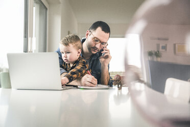 Busy father working at table in home office with son sitting on his lap - UUF16867