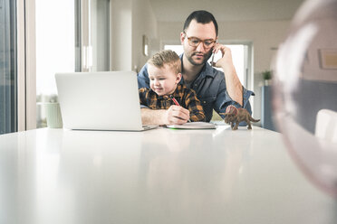 Beschäftigter Vater arbeitet am Tisch im Heimbüro mit seinem Sohn auf dem Schoß - UUF16869