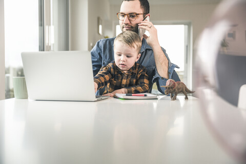 Father working at table in home office with son sitting on his lap stock photo