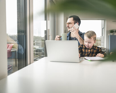 Vater bei der Arbeit am Tisch im Heimbüro mit seinem Sohn auf dem Schoß - UUF16872