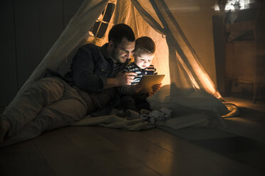Vater und Sohn teilen sich ein Tablet in einem dunklen Zelt zu Hause - UUF16878