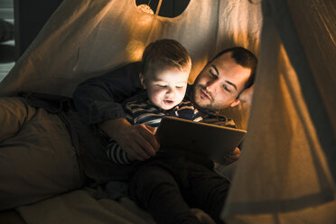 Father and son sharing a tablet in a dark tent at home - UUF16881