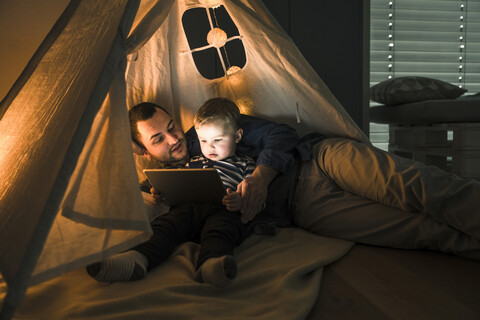 Vater und Sohn teilen sich ein Tablet in einem dunklen Zelt zu Hause, lizenzfreies Stockfoto