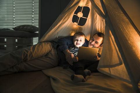 Vater und Sohn teilen sich ein Tablet in einem dunklen Zelt zu Hause, lizenzfreies Stockfoto