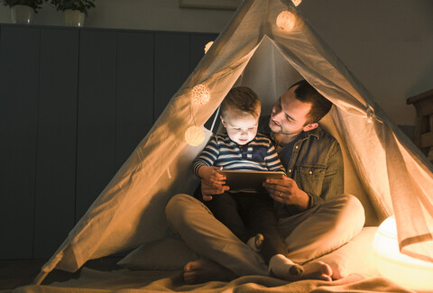 Father and son sharing a tablet in a dark tent at home stock photo