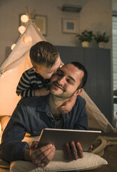 Son kissing father with tablet at an illuminated tent at home - UUF16891