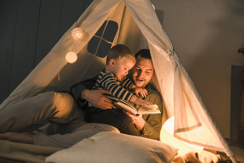 Father reading book to son at an illuminated tent at home - UUF16892