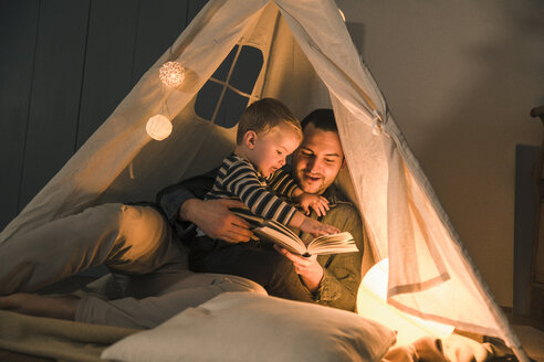 Vater liest seinem Sohn in einem beleuchteten Zelt zu Hause ein Buch vor - UUF16892