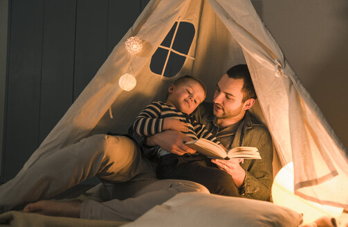 Vater liest seinem Sohn in einem beleuchteten Zelt zu Hause ein Buch vor - UUF16893