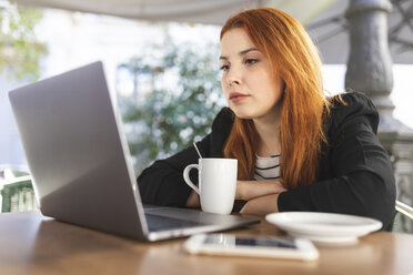 Porträt einer rothaarigen jungen Frau in einem Straßencafé mit Blick auf einen Laptop - WPEF01442