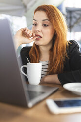 Porträt einer rothaarigen jungen Frau mit Nasenpiercing vor einem Laptop in einem Straßencafé - WPEF01444