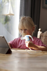 Porträt eines blonden kleinen Mädchens, das einen Asthma-Inhalator benutzt und auf ein digitales Tablet schaut - GAF00100