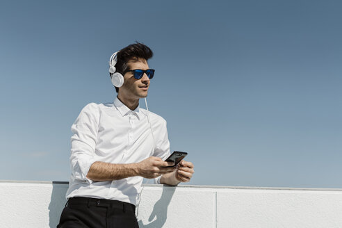 Porträt eines Geschäftsmannes, der mit Kopfhörern und Smartphone auf der Dachterrasse Musik hört - AFVF02668