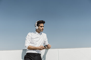 Porträt eines Geschäftsmannes, der mit Kopfhörern und Smartphone auf der Dachterrasse Musik hört - AFVF02671