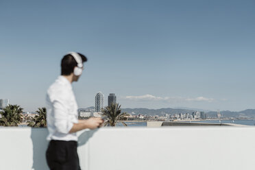 Spanien, Barcelona, Geschäftsmann hört Musik mit Kopfhörer und Smartphone auf Dachterrasse mit Blick in die Ferne - AFVF02676