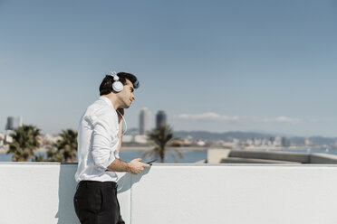 Spanien, Barcelona, Geschäftsmann hört Musik mit Kopfhörern und Smartphone auf der Dachterrasse - AFVF02677