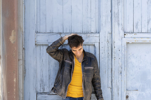 Smiling man wearing leather jacket standing in front of lightblue wooden door - AFVF02702