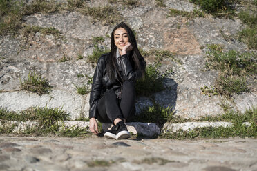 Portrait of smiling young woman sitting on curb - GIOF05963