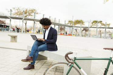 Spain, Barcelona, businessman with bicycle in the city sitting on bench using laptop - VABF02273