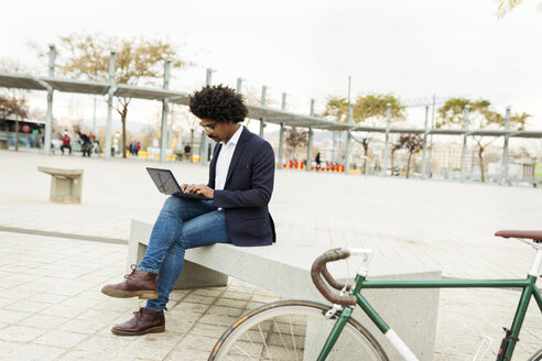 Spanien, Barcelona, Geschäftsmann mit Fahrrad in der Stadt, der auf einer Bank sitzt und einen Laptop benutzt - VABF02273