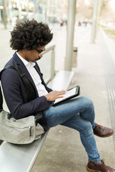 Spain, Barcelona, businessman in the city sitting on bench at a station using tablet - VABF02297