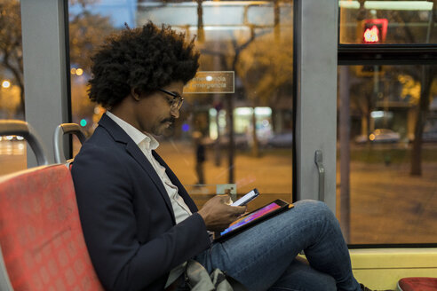 Spanien, Barcelona, Geschäftsmann in einer nächtlichen Straßenbahn mit Handy und Tablet - VABF02323