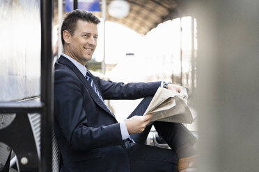 Smiling businessman reading newspaper on station platform - DIGF06424