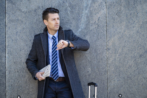 Geschäftsmann mit Koffer, der an einer Wand steht und die Zeit überprüft - DIGF06460