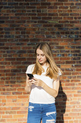 Junge Frau vor einer Backsteinmauer, mit Smartphone - GIOF06051