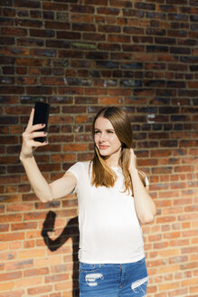 Junge Frau vor einer Backsteinmauer, die ein Selfie mit ihrem Smartphone macht - GIOF06052