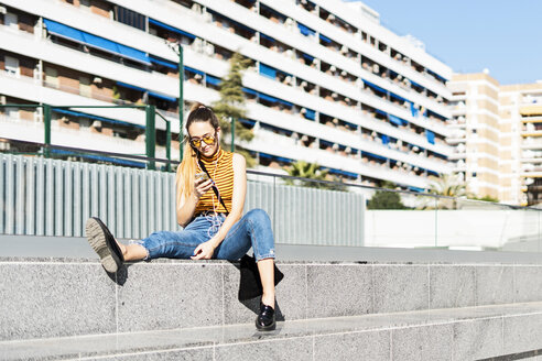 Spanien, junges Mädchen mit Smartphone und Kopfhörern auf einer Treppe in der Stadt sitzend - ERRF00860