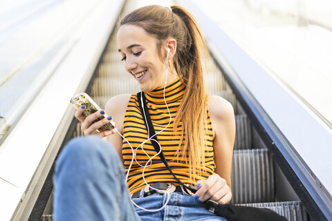 Teenager-Mädchen sitzt mit Kopfhörern und Smartphone auf der Rolltreppe, lizenzfreies Stockfoto