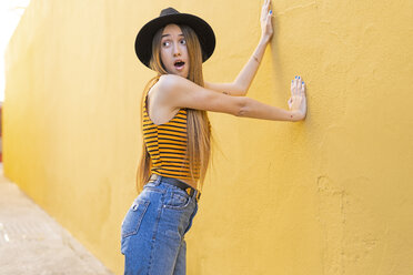Portrait of surprised teenage girl wearing hat at yellow wall - ERRF00886