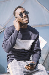 Spain, Madrid, portrait of smiling young man with smartphone wearing mirrored sunglasses putting on earphones - OCMF00340