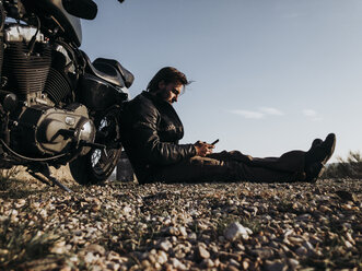 Mann mit Custum-Motorrad macht Pause, sitzt auf dem Boden und telefoniert - OCMF00342