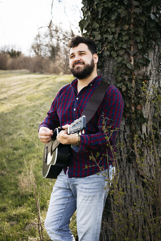 Mann spielt Gitarre an einem Baum auf einer Wiese, lizenzfreies Stockfoto