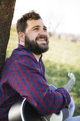 Porträt eines lächelnden Mannes mit Gitarre an einem Baum auf einer Wiese - HMEF00278