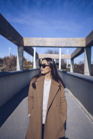 Junge Frau mit Mantel und Sonnenbrille steht auf einer Brücke, lizenzfreies Stockfoto