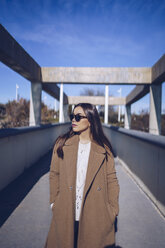 Young woman wearing coat and sunglasses standing on a bridge - RSGF00168