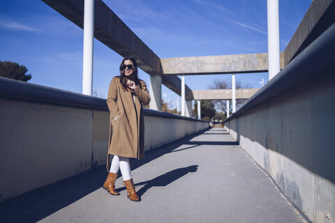 Lächelnde junge Frau mit Mantel und Sonnenbrille auf einer Brücke, lizenzfreies Stockfoto
