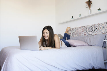 Portrait of smiling young woman lying on bed using laptop - IGGF00927
