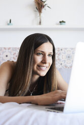 Porträt einer lächelnden jungen Frau, die auf dem Bett liegt und einen Laptop benutzt - IGGF00929