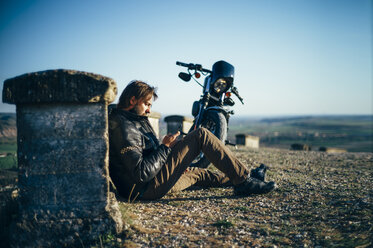 Mann mit Custum-Motorrad macht Pause, sitzt auf dem Boden und telefoniert - OCMF00344