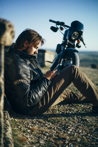 Mann mit Custum-Motorrad macht Pause, sitzt auf dem Boden und telefoniert, lizenzfreies Stockfoto