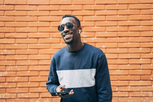 Portrait of happy man wearing sunglasses listening music with wireless earphones and smartphone - OCMF00348