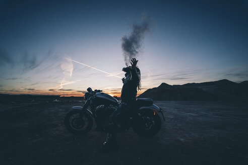 Bärtiger Mann mit Dreadlocks sitzt bei Sonnenuntergang auf einem Motorrad und raucht eine elektronische Zigarette - OCMF00352