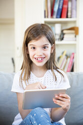Portrait of happy little girl sitting on the couch at home using digital tablet - LVF07956