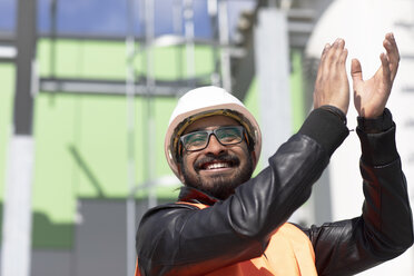 Porträt eines glücklichen Bauingenieurs vor einem Kraftwerk mit Schutzhelm und Sicherheitsweste, der in die Hände klatscht - SGF02344