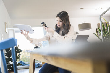 Young woman sitting at table at home holding smartphone and plane model - UUF16950