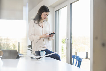 Junge Frau am Tisch zu Hause, die auf ihr Smartphone schaut - UUF16953